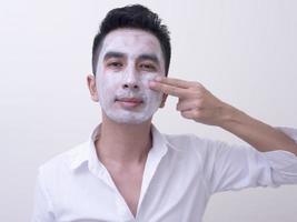 jovem bonito asiático aplicando creme no rosto com carinha sorridente, conceito de cuidados com a pele foto