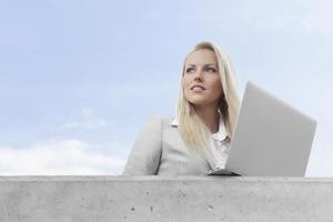 jovem empresária com laptop, olhando para longe no terraço contra o céu foto