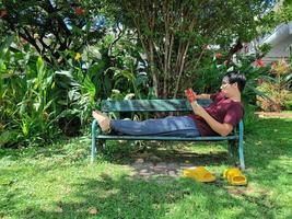 jovem asiático de óculos com cabelo preto, segurando um livro sentado com as pernas esticadas em um banco. feliz e relaxe. o que estão pensando na cidade do parque de verão foto