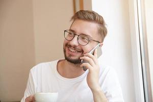 retrato de jovem bonito sorrindo largamente, segurando uma xícara de café com uma mão e telefone na outra, estando de bom humor, usando óculos e camiseta branca foto