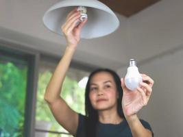 mulher asiática trocando a lâmpada em sua casa. substitua a lâmpada fluorescente por led, mostrando a lâmpada led para a câmera.. foco seletivo. conceito de economia de energia. foto