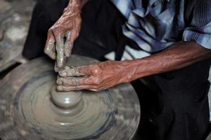 seletiva focada nas mãos sujas de pele enrugada do velho moldando o trabalho de barro na roda de fiar para fazer o jarro tradicional foto