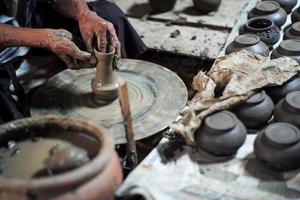 seletivo focado nas mãos sujas de pele enrugada do velho moldando o trabalho de barro na roda de fiar para fazer o pote com grupo borrado de potes de barro em primeiro plano foto