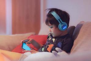 retrato noturno cinematográfico jovem usando fone de ouvido para jogar online na internet com amigo, garoto sentado no sofá assistindo desenho animado no tablet criança relaxando em casa no fim de semana foto