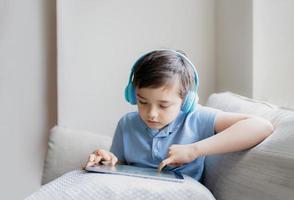 menino feliz usando fone de ouvido para jogar na internet com amigos, criança sentada ao lado da janela lendo ou assistindo desenhos animados no tablet, criança da escola fazendo lição de casa online em casa. foto