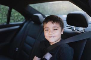 garoto de escola feliz sentado no assento de carro de segurança olhando com rosto sorridente, criança positiva sentada no banco do passageiro de trás com um cinto de segurança, menino viajando para a escola de carro. de volta à escola foto