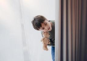 menino bonito segurando ursinho de pelúcia atrás da cortina de renda com luz brilhante na manhã, criança feliz com sorriso no peitoril da janela e brinca de esconde-esconde atrás da cortina foto