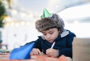 menino bonito com chapéu de festa usando caneta rosa desenhando ou escrevendo na cor do papel com fundo claro embaçado, criança se divertindo na festa de aniversário, criança fazendo atividade no feriado de natal ou ano novo foto