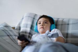 retrato de criança feliz usando fones de ouvido e olhando para cima assistindo tv, criança sentada no sofá segurando o controle remoto, menino feliz ouvindo música relaxante na sala de estar em casa foto