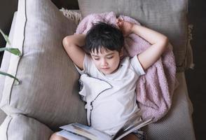 garoto de escola feliz deitado no sofá lendo um livro com a luz da manhã brilhando da janela. vista superior menino lendo história relaxando em casa no fim de semana, conceito positivo de crianças foto