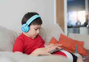 menino feliz usando fone de ouvido para jogar na internet com amigos, criança sentada no sofá lendo ou assistindo desenhos animados no tablet, garoto da escola fazendo lição de casa online em casa. foto