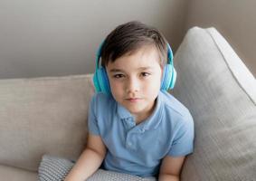 menino feliz usando fones de ouvido, ouvindo música. lindo garoto olhando para a câmera com rosto sorridente, criança positiva sentada no sofá relaxando no fim de semana na primavera ou verão foto