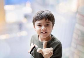 menino feliz olhando para cima com o rosto sorridente segurando o ursinho de pelúcia ao lado do vidro da janela com luz brilhante brilhando, criança positiva brincando com urso marrom de brinquedo macio, criança relaxando em casa de manhã foto