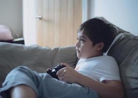 menino feliz jogando videogame on-line com amigos, criança fofa de tiro sincero sentado no sofá segurando o console do jogo. foto