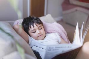 garoto de escola feliz deitado no sofá lendo um livro com a luz da manhã brilhando da janela. menino criança lendo história relaxando em casa no fim de semana, conceito positivo de crianças foto