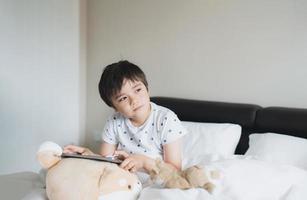 criança jogando jogos online na internet no quarto, retrato de menino usando tablet digitando ou conversando com amigos de manhã, infância sentada na cama fazendo lição de casa, criança da escola com conceito de tecnologia foto