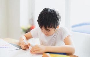 garoto da escola usando caneta de cor desenhando ou escrevendo a carta no papel, menino fazendo lição de casa, criança com caneta escrevendo notas na folha de papel durante a lição aluno bonito fazendo teste, conceito de educação em casa foto