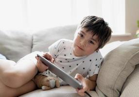 jovem de retrato de alta chave jogando no tablet, criança jogando na internet, criança de retrato cinematográfico fazendo lição de casa on-line em casa, criança sentada no sofá com luz brilhando da janela. foto