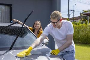 jovem casal caucasiano está sorrindo enquanto usa esponja e água com sabão para lavar o carro na garagem em casa foto