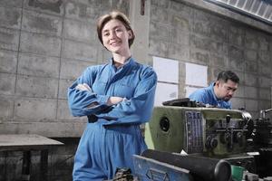 retrato de jovem trabalhadora da indústria feminina branca fica, braços cruzados, olha para a câmera e sorri, parceiro masculino trabalha com máquina atrás dela na fábrica de fabricação, engenheiros mecânicos profissionais. foto