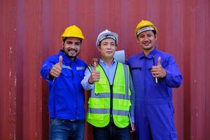 retrato de três trabalhadores da indústria asiática com uniforme de segurança e capacetes no fundo da folha de metal, polegar para cima, olhe para a câmera e sorria, transporte de carga, negócios profissionais de transporte de mercadorias. foto