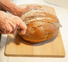 as mãos da mulher idosa cortam pão de centeio redondo em farinha na placa da cozinha foto