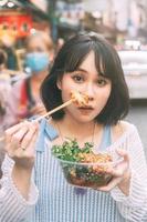 jovem mulher asiática adulta comendo comida de lula grelhada picante no mercado de chinatown do sudeste asiático foto