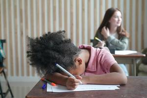 criança pré-escolar desenhando com lápis de cor em papel branco na mesa em sala de aula com amigos foto