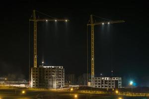 guindastes de torre e edifícios altos de vários andares inacabados em construção à noite no canteiro de obras iluminado foto