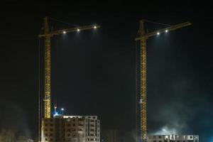 guindastes de torre e edifícios altos de vários andares inacabados em construção à noite no canteiro de obras iluminado foto