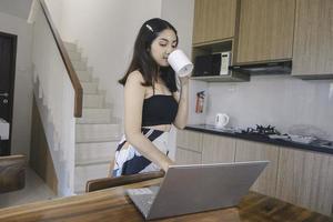 jovem mulher asiática passa seu tempo em casa trabalhando remotamente na sala de jantar tendo uma conferência online em seu laptop enquanto carrega uma caneca. foto