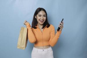 retrato asiático feliz bela jovem em pé animado segurando a sacola de compras on-line e smartphone na mão, estúdio tiro isolado em fundo azul foto