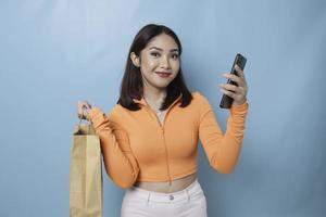 retrato asiático feliz bela jovem em pé animado segurando a sacola de compras on-line e smartphone na mão, estúdio tiro isolado em fundo azul foto