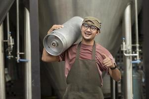 jovem de avental de couro segurando barril de cerveja na cervejaria moderna, trabalhador de cervejaria artesanal foto