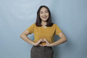 mulher asiática jovem e atraente se sente feliz e romântico formas gesto de coração expressa sentimentos ternos veste casual t-shirt amarela contra fundo azul. conceito de carinho e cuidado de pessoas foto