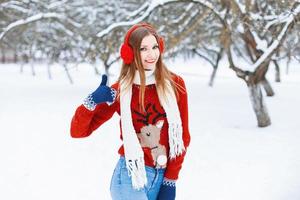 linda jovem de suéter vermelho com fones de ouvido de rena e inverno para se divertir no parque. mostrando os polegares para cima sinal de qualidade foto