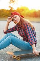 menina alegre em uma camisa e um boné de beisebol, sentado no asfalto com um pôr do sol skateboard.beautiful. foto