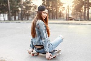 jovem linda na moda em roupas elegantes, sentado em um skate ao pôr do sol foto