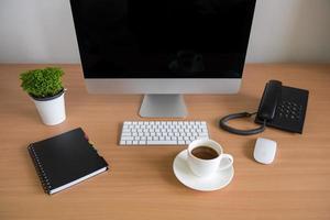 mesa de escritório com computador pessoal, caderno, telefone, teclado, mouse, xícara de café e vaso de plantas foto