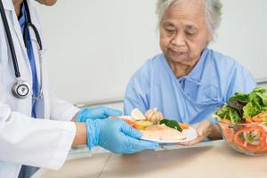 paciente idosa asiática ou idosa tomando café da manhã e comida saudável vegetal com esperança e feliz enquanto está sentado e com fome na cama no hospital. foto