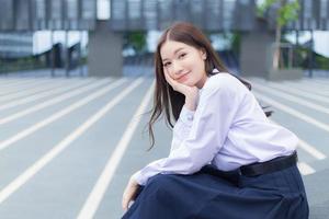 linda estudante asiática do ensino médio no uniforme escolar com aparelho nos dentes senta e sorri com confiança enquanto ela olha para a câmera alegremente com o prédio ao fundo. foto