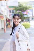 confiante jovem mulher asiática que veste uma camisa branca e uma bolsa de ombro sorri alegremente enquanto ela está caminhando para trabalhar no escritório na cidade. foto