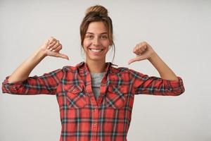 alegre jovem morena bonita de pé sobre fundo branco com polegares levantados, apontando para si mesma com um sorriso feliz, vestindo camisa quadriculada foto