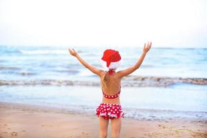 Menina de 5 anos de maiô vermelho e chapéu de papai noel na praia está feliz, grita, pula e acena as mãos com felicidade. natal e ano novo passeios de inverno para países quentes. turismo para férias nos trópicos foto