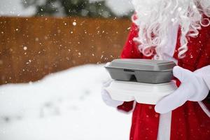 recipientes de serviço de entrega de comida nas mãos do papai noel ao ar livre na neve. promoção de véspera de natal. pedido quente pronto, caixa plástica descartável. catering de férias de ano novo. copie o espaço, simular foto