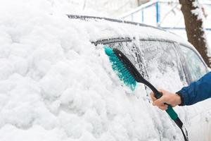 um homem escova a neve de um carro após uma nevasca. uma mão em uma jaqueta azul com uma vassoura de carro no corpo branco. condições meteorológicas de inverno foto