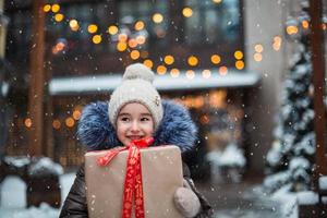 retrato de menina alegre com uma caixa de presente para o natal em uma rua da cidade no inverno com neve em um mercado festivo com enfeites e luzes de fadas. agasalhos, gorro de malha, cachecol e peles. ano Novo foto