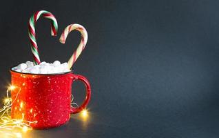 caneca vermelha com marshmallows e pirulito de caramelo em forma de coração e luzes de guirlandas em um fundo preto. natal, ano novo, clima festivo. espaço de cópia foto