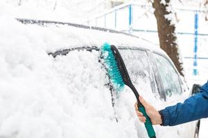 um homem escova a neve de um carro após uma nevasca. uma mão em uma jaqueta azul com uma vassoura de carro no corpo branco. condições meteorológicas de inverno foto