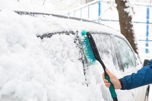 um homem escova a neve de um carro após uma nevasca. uma mão em uma jaqueta azul com uma vassoura de carro no corpo branco. condições meteorológicas de inverno foto